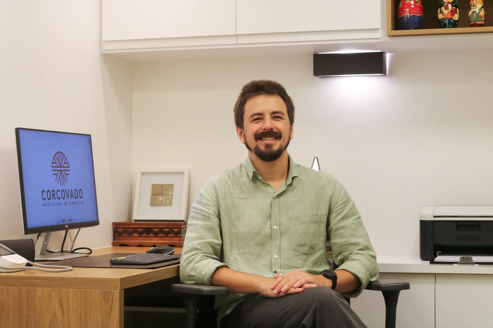 João sentado em seu consultório, ao lado da mesa e do computador